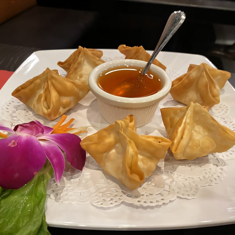 Golden fried Thai dumplings with sweet chili dipping sauce and an orchid garnish on a white plate.