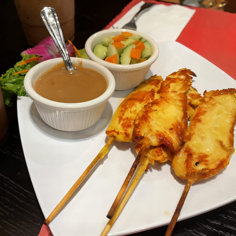 Thai chicken satay skewers with peanut sauce and cucumber salad on a white plate.