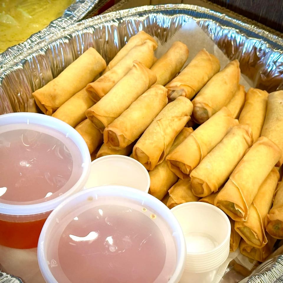 A catering tray filled with golden-brown Thai spring rolls and two containers of dipping sauce.