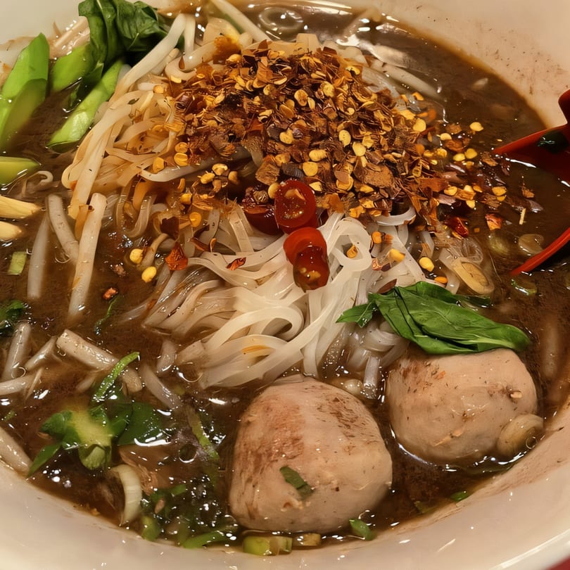 Close-up of Thai boat noodles with meatballs, bean sprouts, greens, and chili flakes.