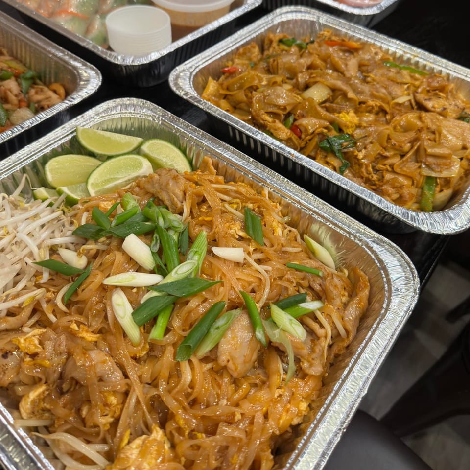 Multiple aluminum containers of Thai food, including Pad See Ew and Pad Thai with lime wedges.