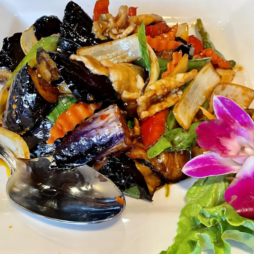 A Thai eggplant dish with mushrooms, bell peppers, and basil, garnished with an orchid.